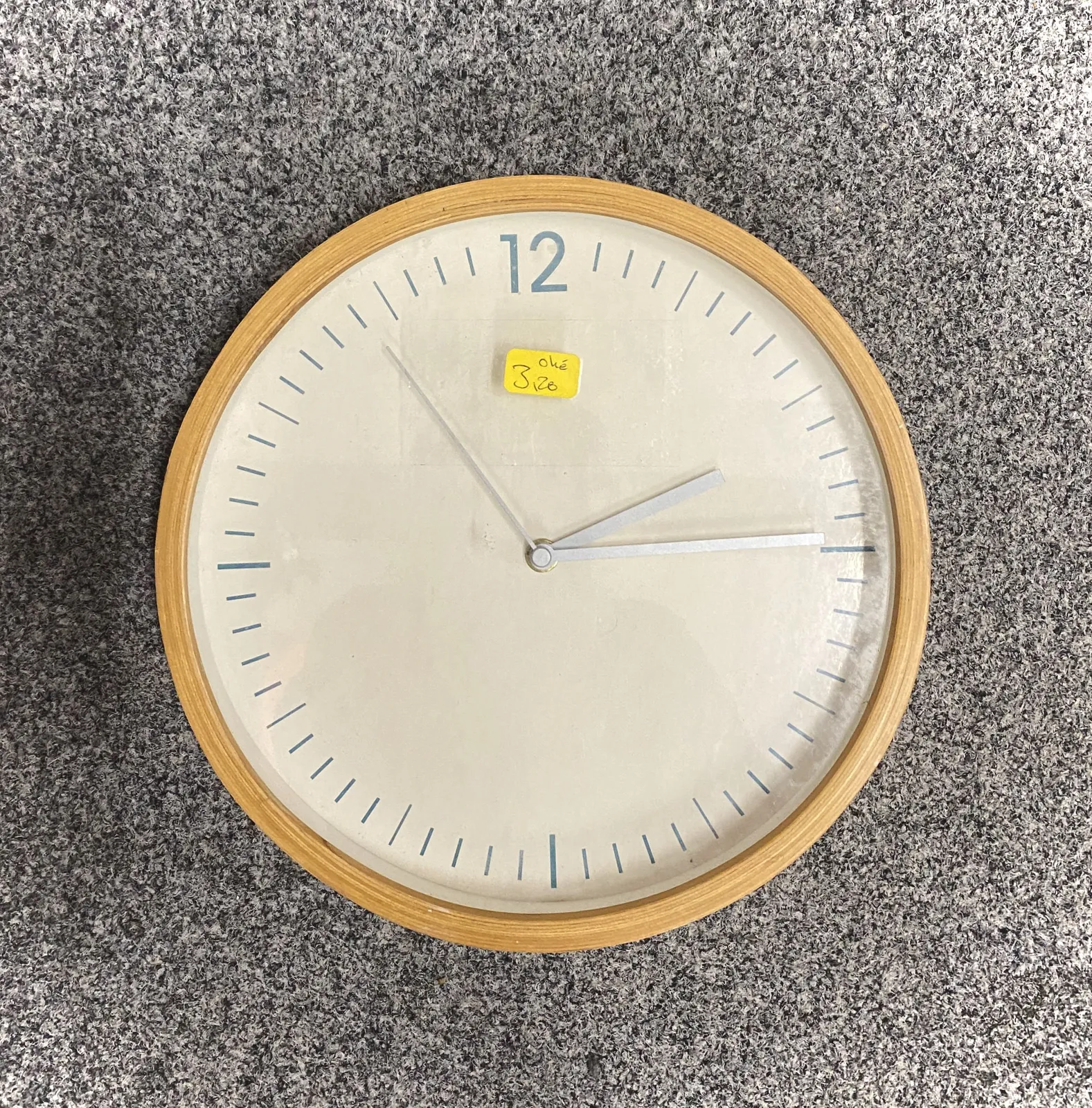 A simple, modern wall clock. Only the 12, at the top, is displayed as a number, the rest of the indicators are small dashes. There’s a small yellow sticker on it that says Okay, and a price of 3,20. The clock points at 14 minutes past 2.