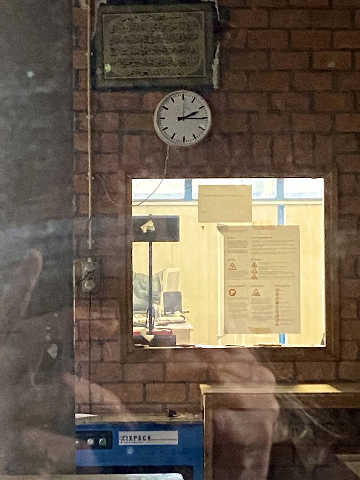 A clock attached to a brick wall in some kind of workshop. It points at a quarter past 2.