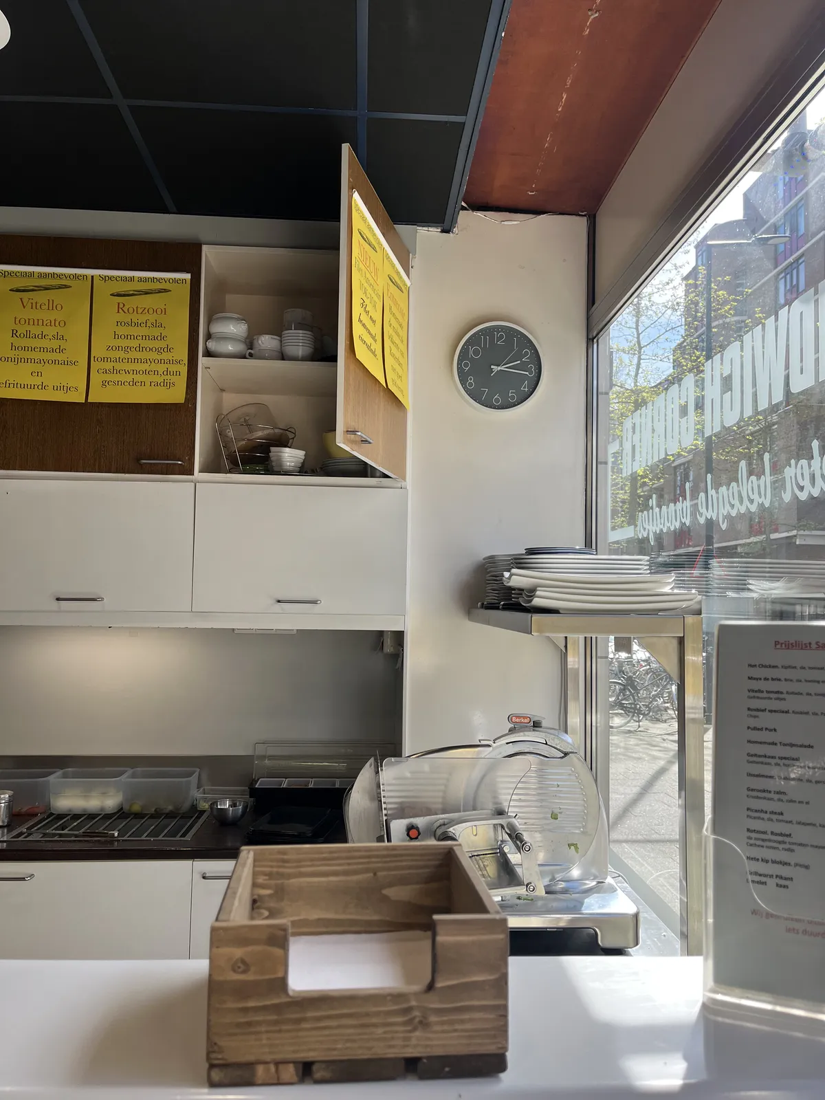A clock on a wall, between the window and some cupboards, in a sandwich shop. It points at 16 minutes past two.