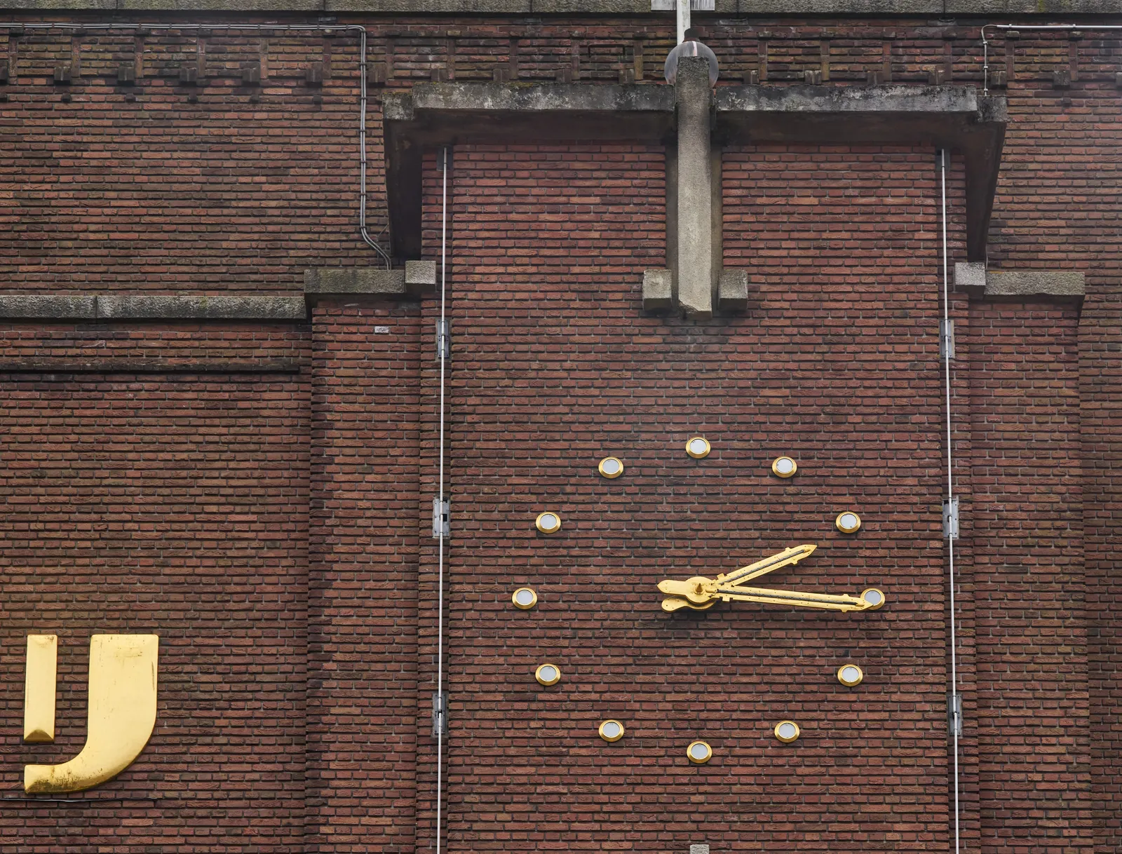 On a monumental, rather old brick wall there are some large golden letters that spell IJ, and a golden clock that points at 16 past two.
