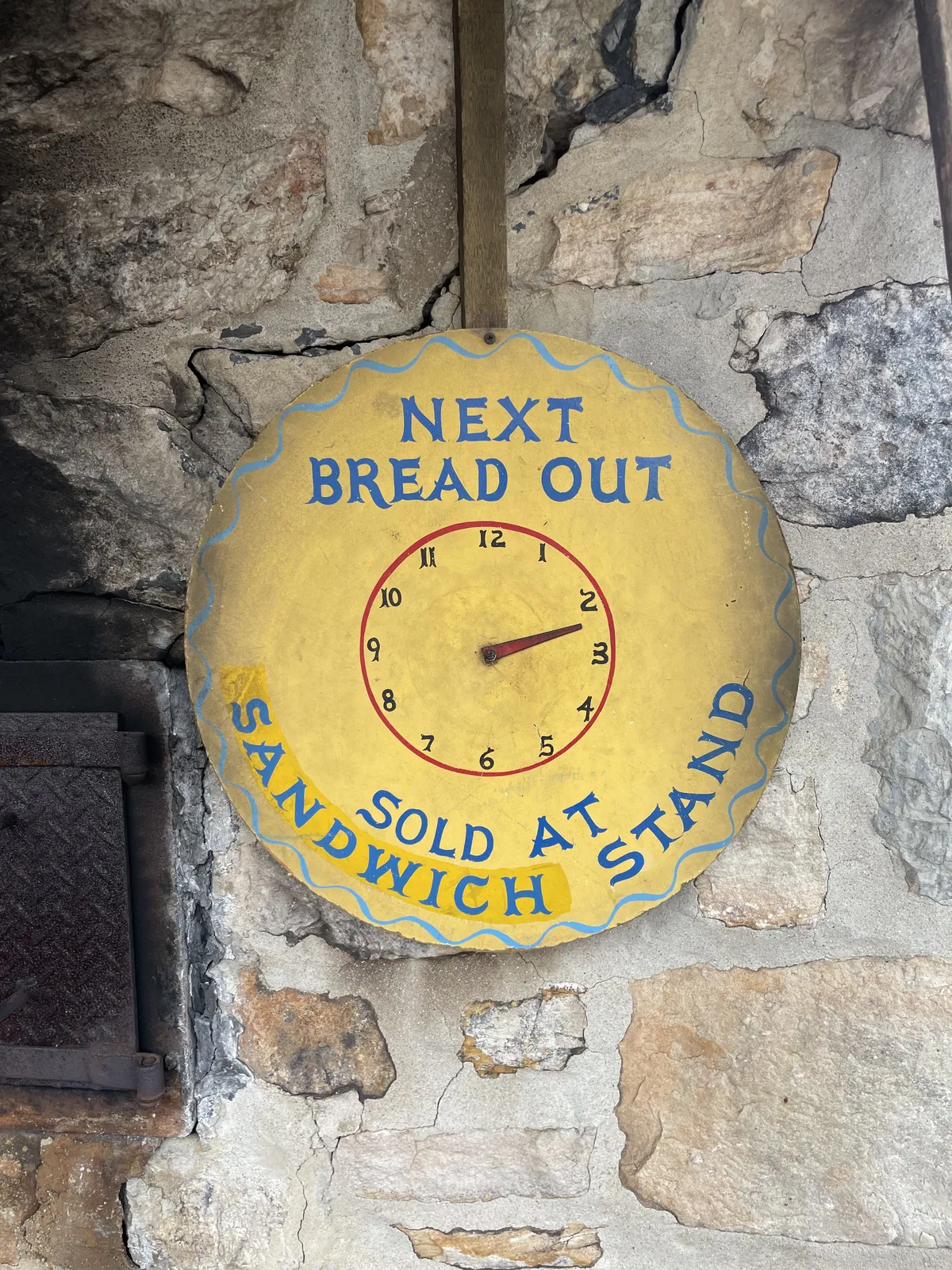 On a stone wall there’s a clock with one, red hand. Painted on the clock is the message, in blue, that the next bread out is sold at the sandwich stand. Since it’s just a single hand it is a bit hard to see what at time exactly, but it seems like the next bread will be out at approximately 20 minutes past 2.
