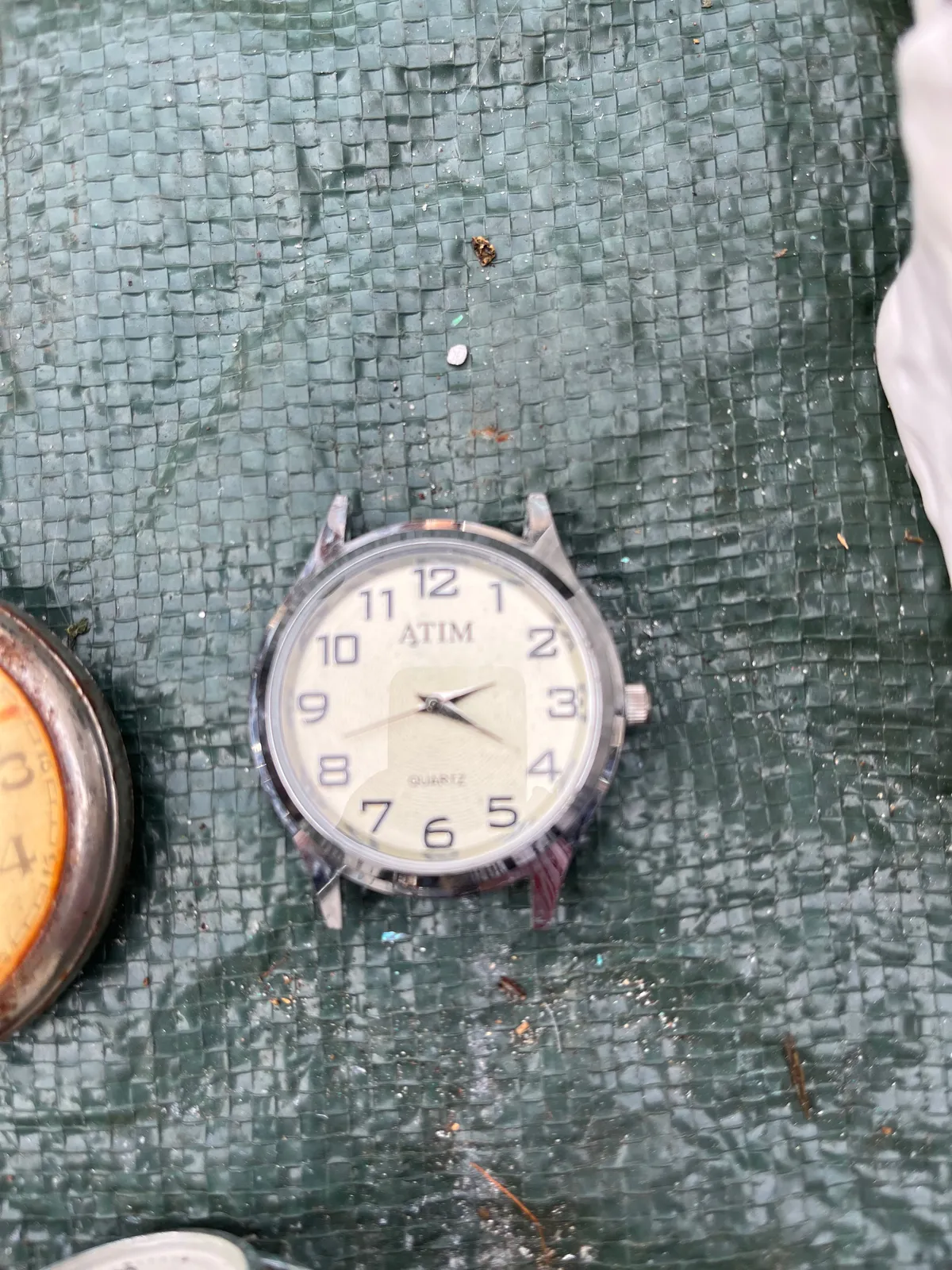 A watch without straps with the word ATIM printed on its face. It points at 20 past 2.