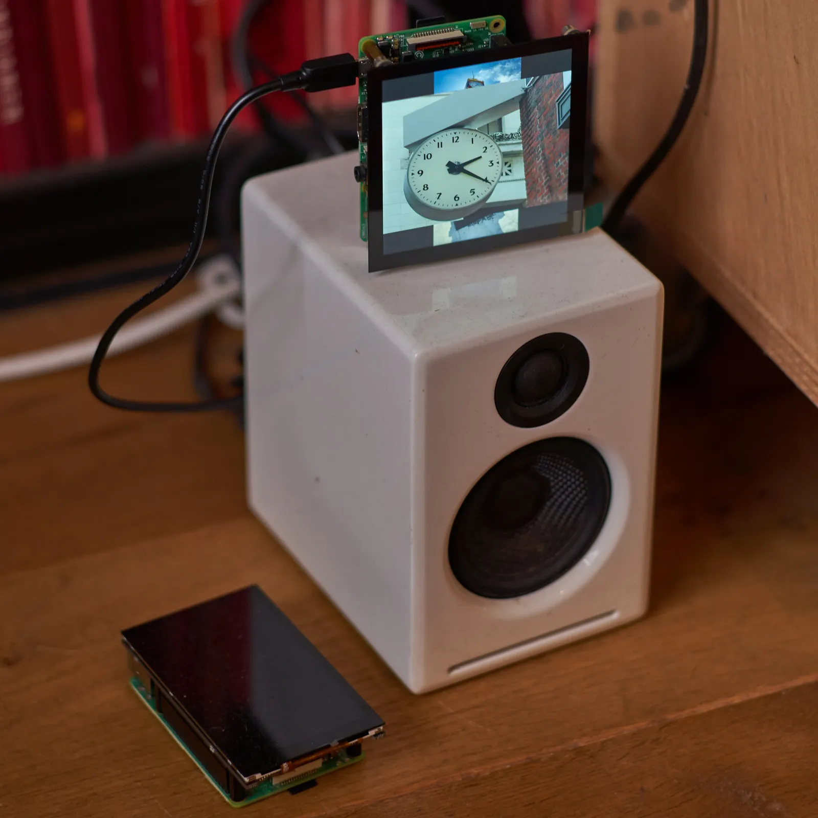 A little square screen with a photo of a clock on it (it points at 21 minutes past two). The screen is placed on top of a small speaker. There’s a second, non working screen laying on the ground next to it.