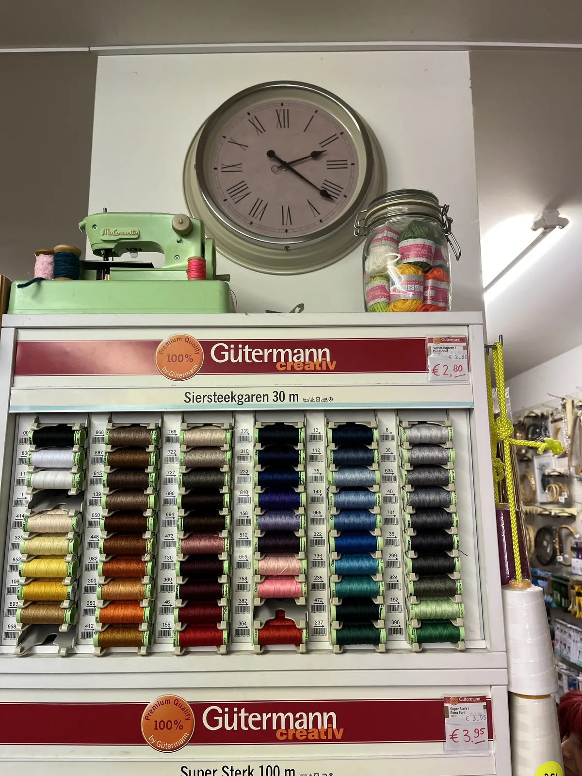 Above a display filled with spools with colourful yarn, there’s an old sewing machine, and a clock with roman numerals. This clock points at 21 minutes past two.