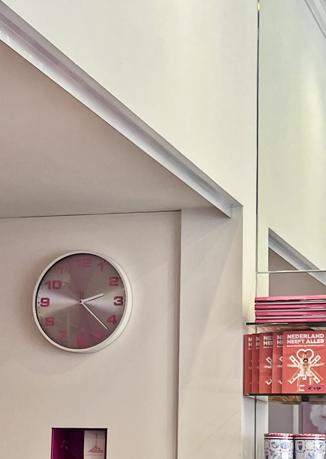 Looking at a recess in a wall. There’s a bookshelf on the right with a book on it, with the title, in Dutch: The Netherlands has everything. On the left there’s a white, silver, magenta clock which points at 22 minutes past 2