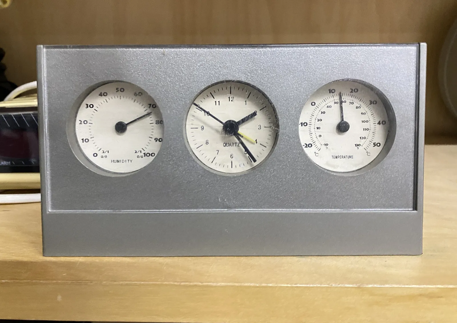 A silver coloured device with three clocks on it. The one on the left shows the humidity, the one on the right the temperature, and the one in the middle is a clock which points at 24 minutes past 2.