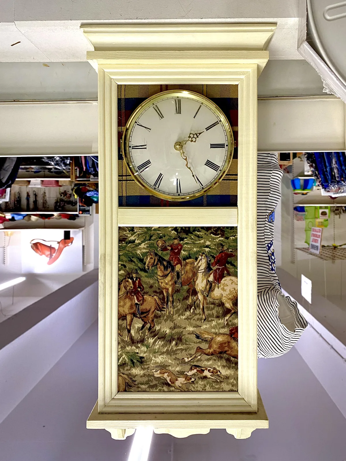 The embroidery on this clock shows a scene with hunters on horseback. There are a few dogs as well. On the clock it is 25 past 2. The clock is placed upside down on the floor. But since the image is rotated, the background is upside down, and the clock looks fine.