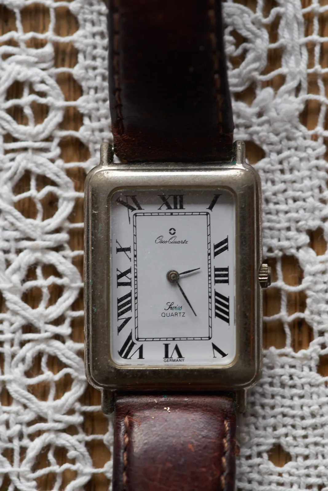 This square watch on a crocheted white fabric background points at 25 minutes past 2.