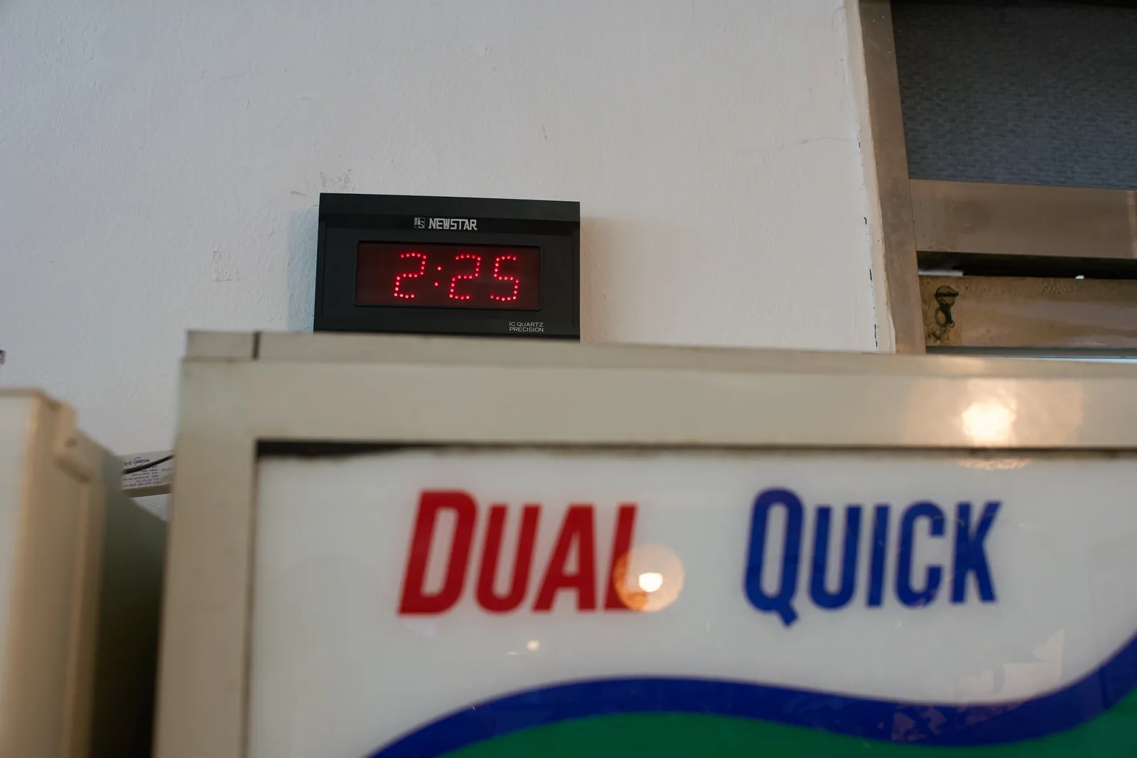 A digital clock on top of a fridge. The fridge has the words Dual Quick on it. The clock says it it 2:25.