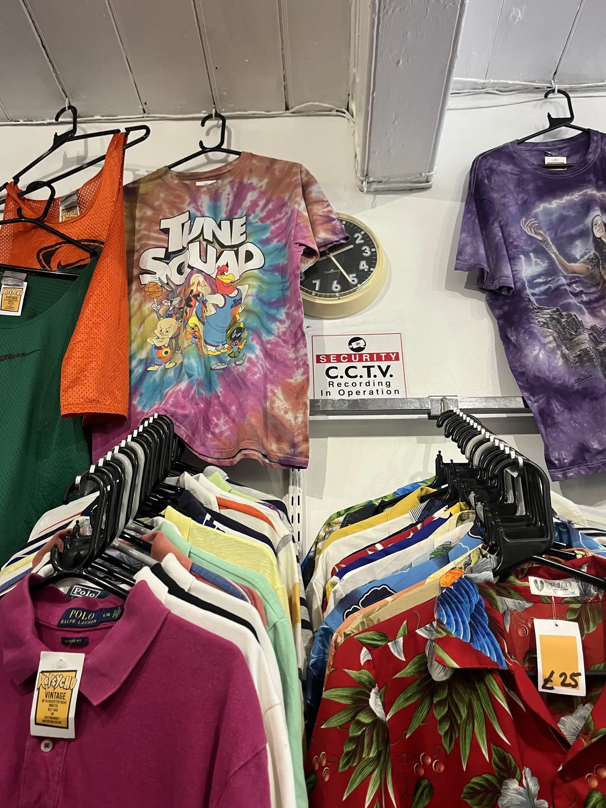 Very many colourful shirts for sale. Half hidden behind some shirts is a wall clock which points at 26 minutes past two.