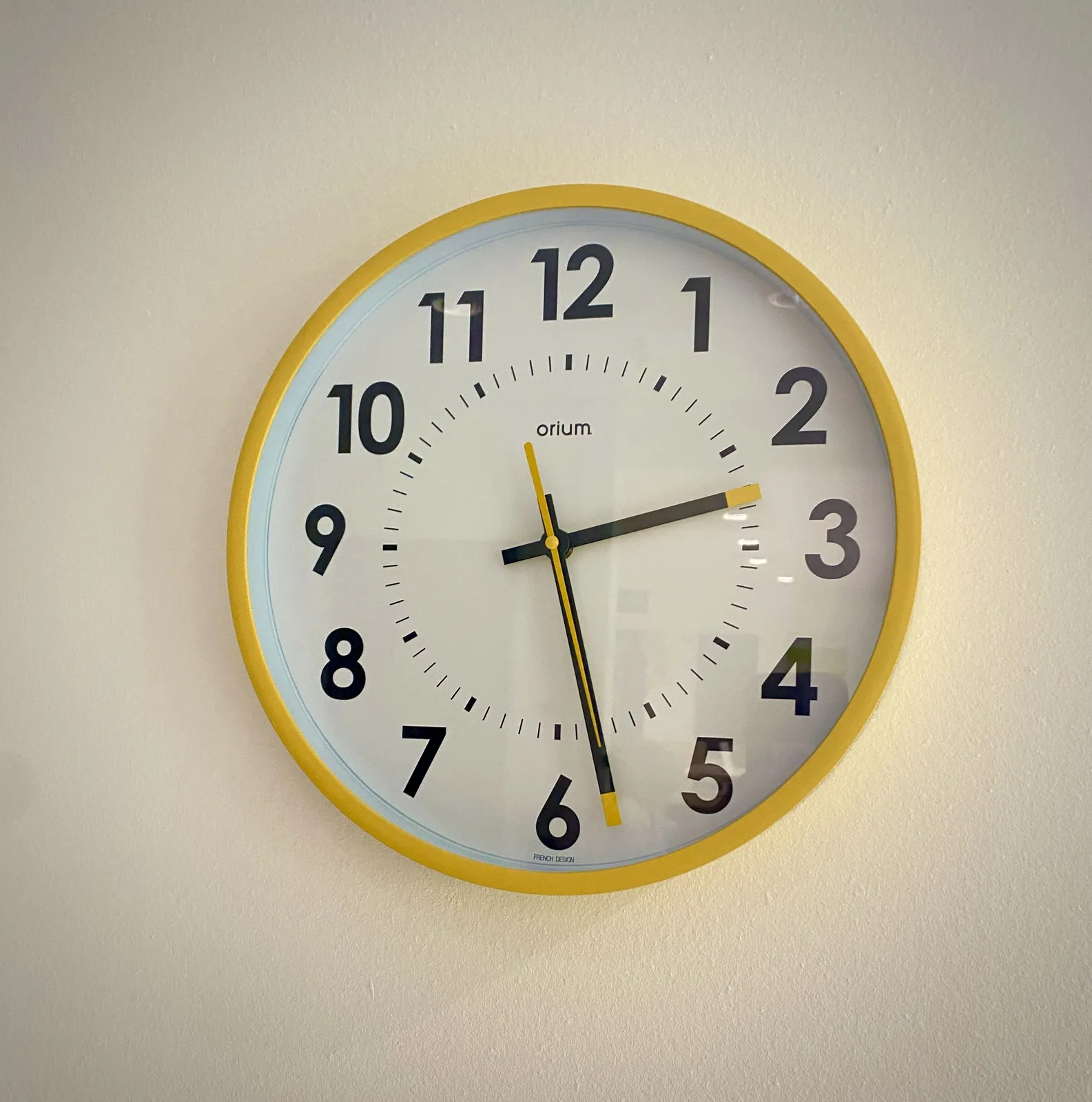 A wall clock with quite a few bright yellow details. For instance, the brim and the endpoints of the hands are yellow. It points at 28 minutes past 2.