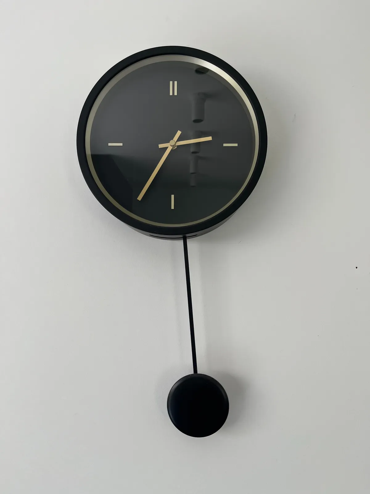 A modern, black clock with a pendulum. It points at 26 minutes to 3.