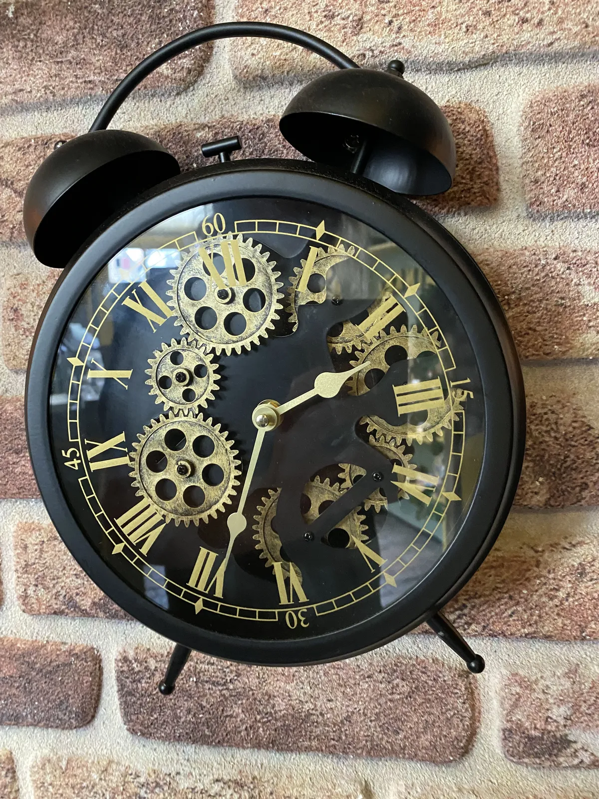 A black and gold clock with many cogs. It’s shaped like an old fashioned alarm clock, but it is a wall clock. It points at 34 minutes past 2.