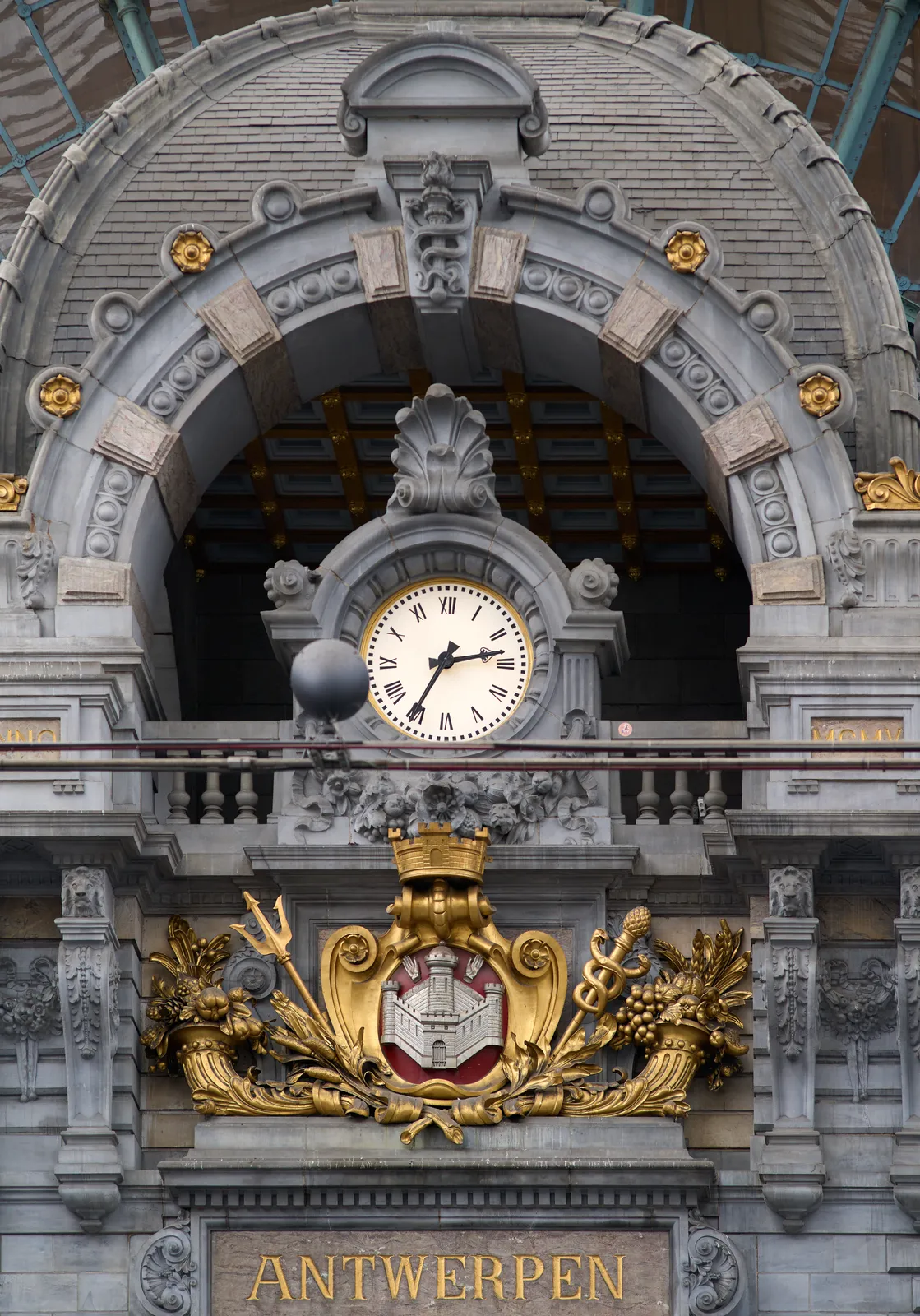 A clock surrounded by gold ornaments, in a grey building, above the word Antwerpen. The clock points at 35 minutes past two.