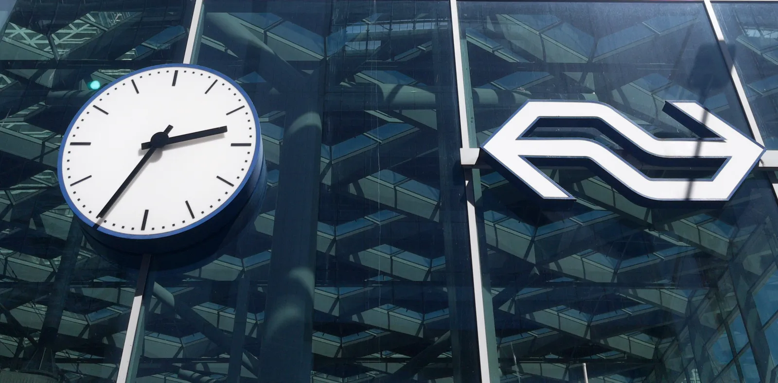 Next to an enormous logo of the Dutch Railways, built into a glass facade, is a large clock, also in the glass facade. It points at 35 past 2