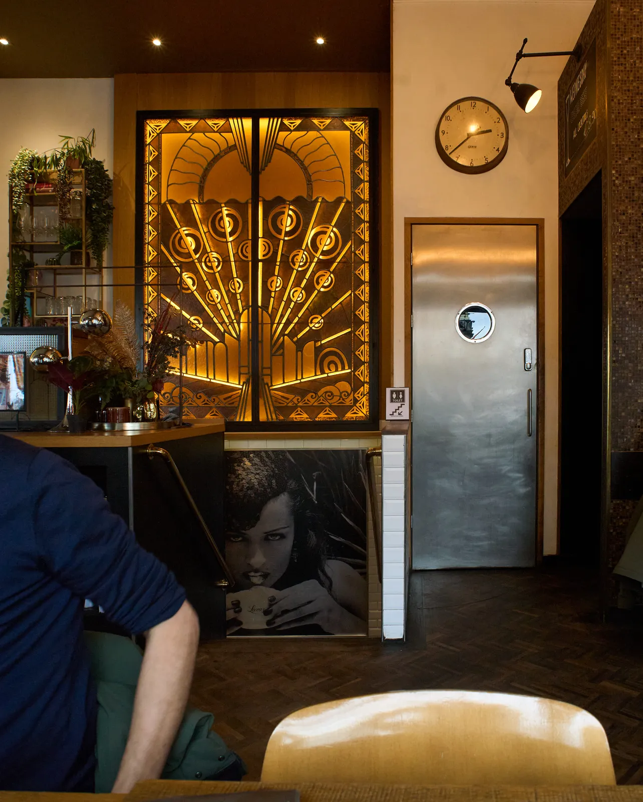 Someone’s arm can be seen, the rest of the body is outside of the picture. In the background lots of ornaments and a door, all in a cafe. There’s also a clock, it points at 22 minutes to 3.