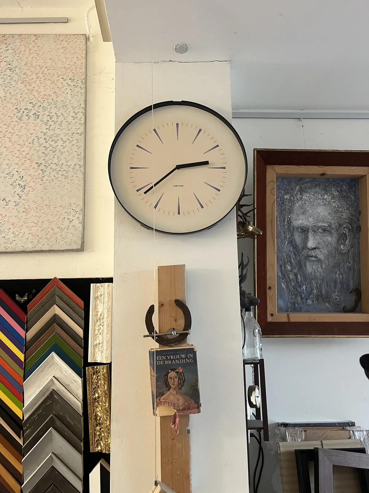 A display of different picture frames, a few works of art, a horseshoe, the cover of a book, and a wall clock, all attached to a wall. The clock has thin pointy hour and minute indicators, and straight, minimalist black hands. It points at 22 minutes to three. 