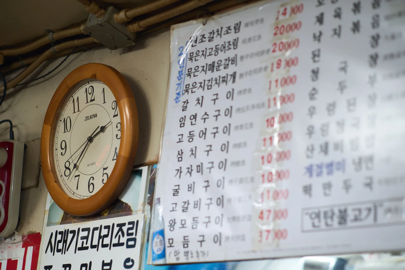 A wall clock, hanging on a wall in a Korean restaurant. There’s a price list on the right that can only be understood if you know Korean. On the face of the clock it says Zelkova Quartz. It points at 38 minutes past 2.