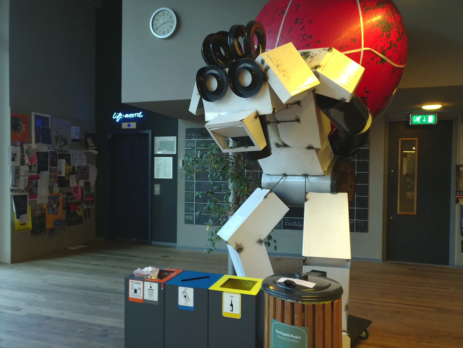 A enormous robot carrying a huge red metal ball stands in front of a few garbage cans. In the background there’s a clock that points at 20 minutes to 3.