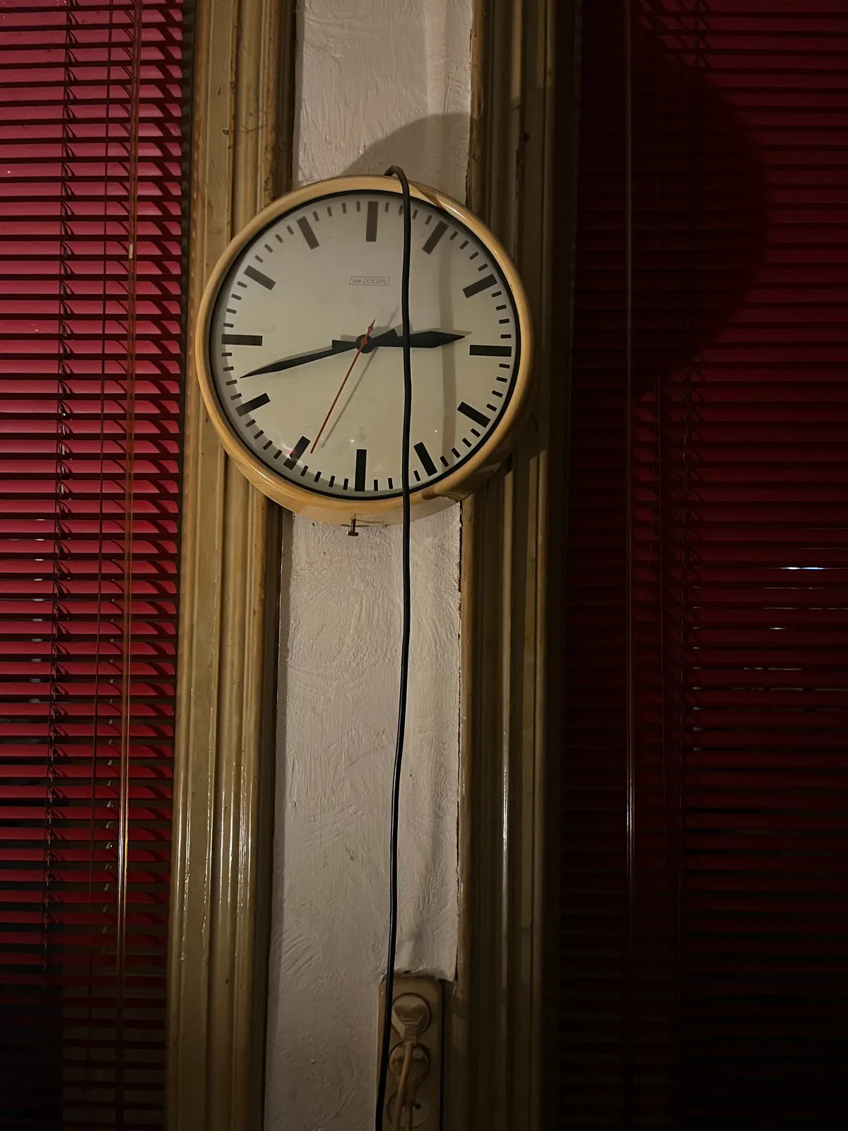 An electric wall clock, hanging on a small wall, between two windows. The windows are covered with red Venetian blinds. The electrical chord hangs over the clock. The clock points at 18 minutes to 3.