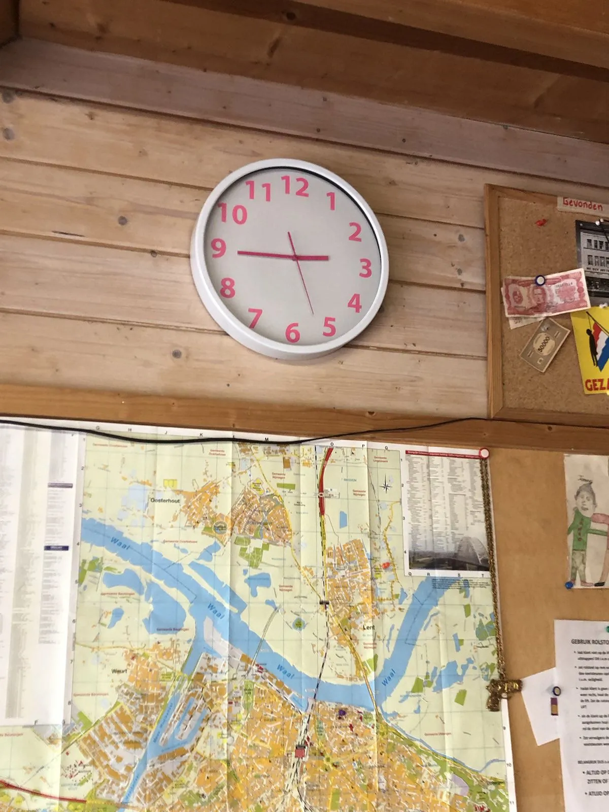 On a wooden wall there’s a bulletin board with a map and some foreign banknotes. There’s a clock on the wall which points at 16 to 3.