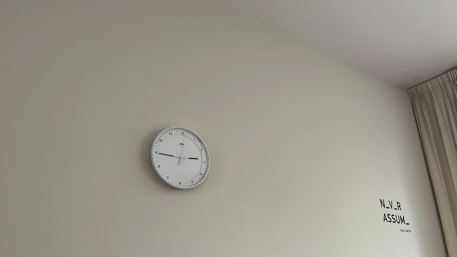 A white wall. In the far corner, on the right, there are white curtains. On the wall a text is printed in black. It says: n_v_r assum_, Paul Smith. There’s also a clock, made by the firma Braun on this wall. It points at a quarter to three.