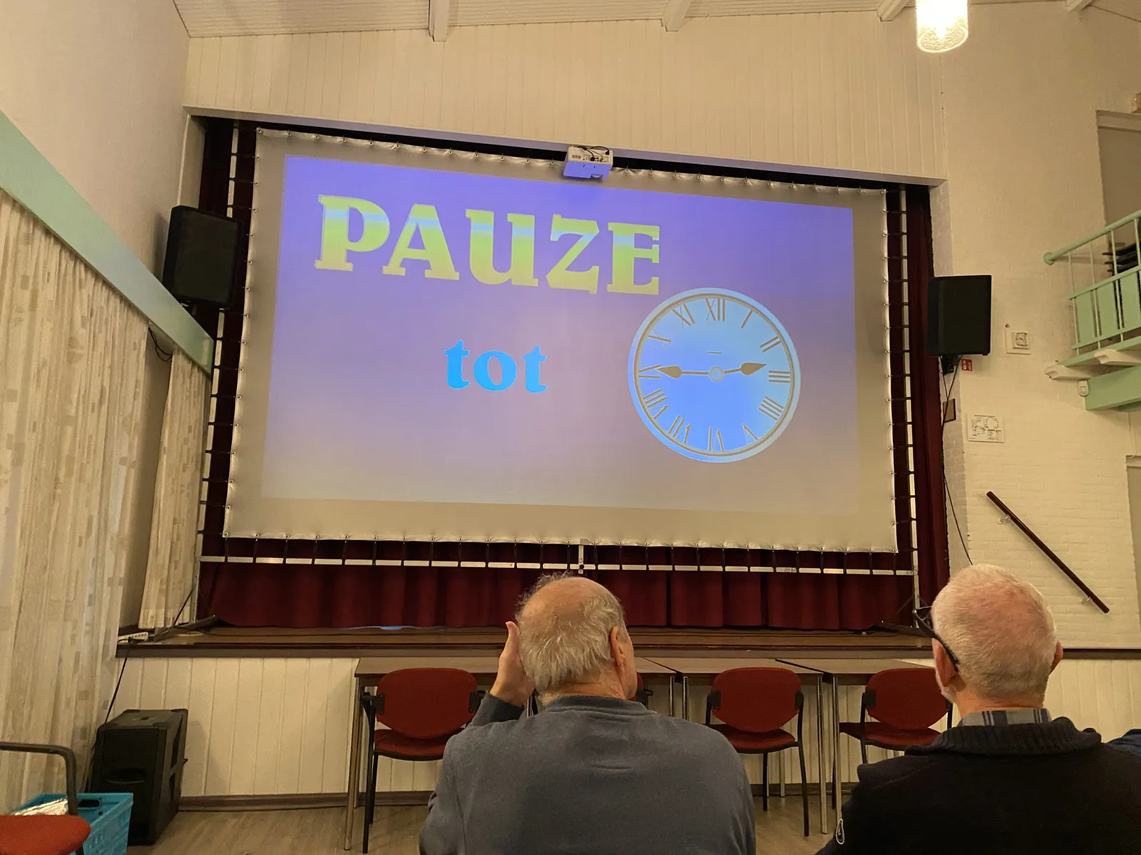 Two old men from behind, looking at a projection with the words A Break Until, and a clock on it. The clock points at a quarter to three.