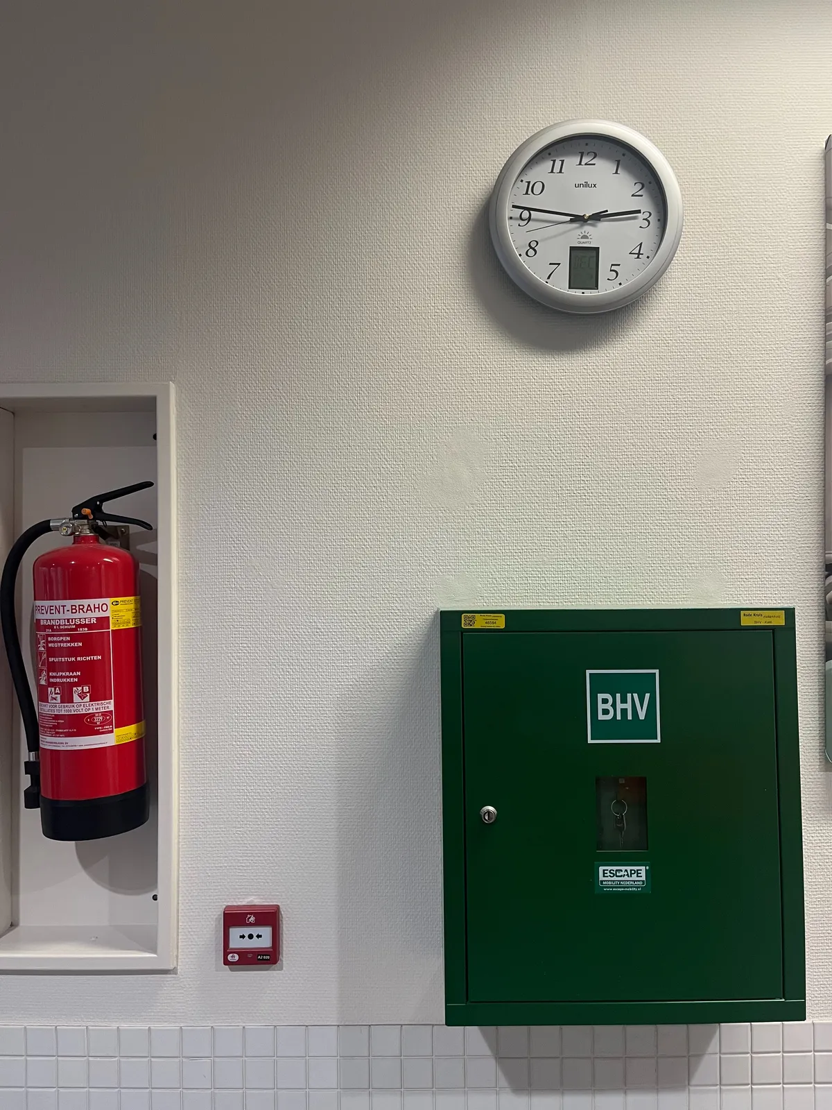 A fire extinguisher, a green cabinet with the letters BHV printed on it, and a clock, all attached to a wall. The clock points at 14 minutes to 3.