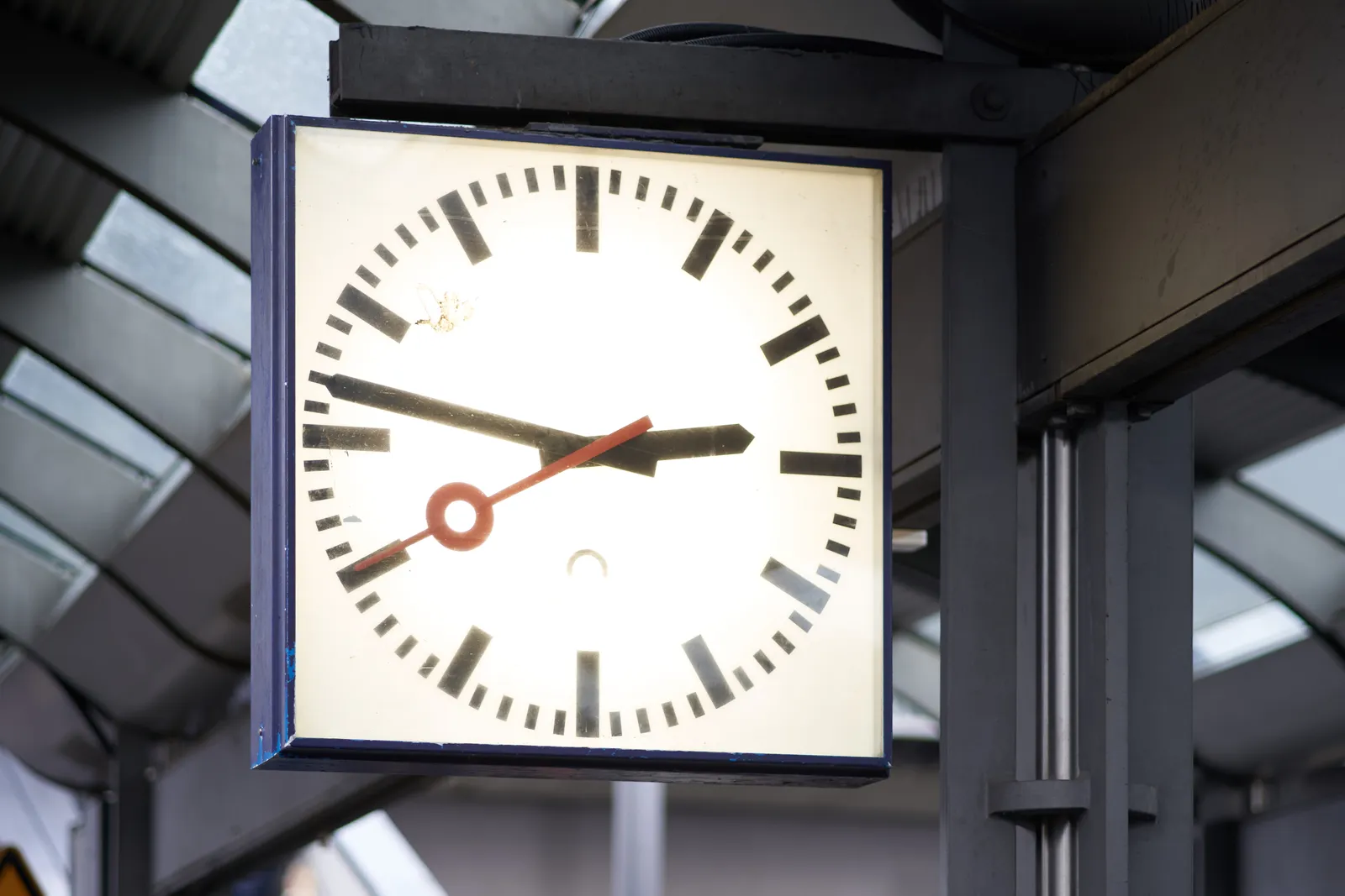 A square modernist clock with black minute indicators and black minute and hour hands. It has a red second hand. The clock points at 13 minutes to 3.