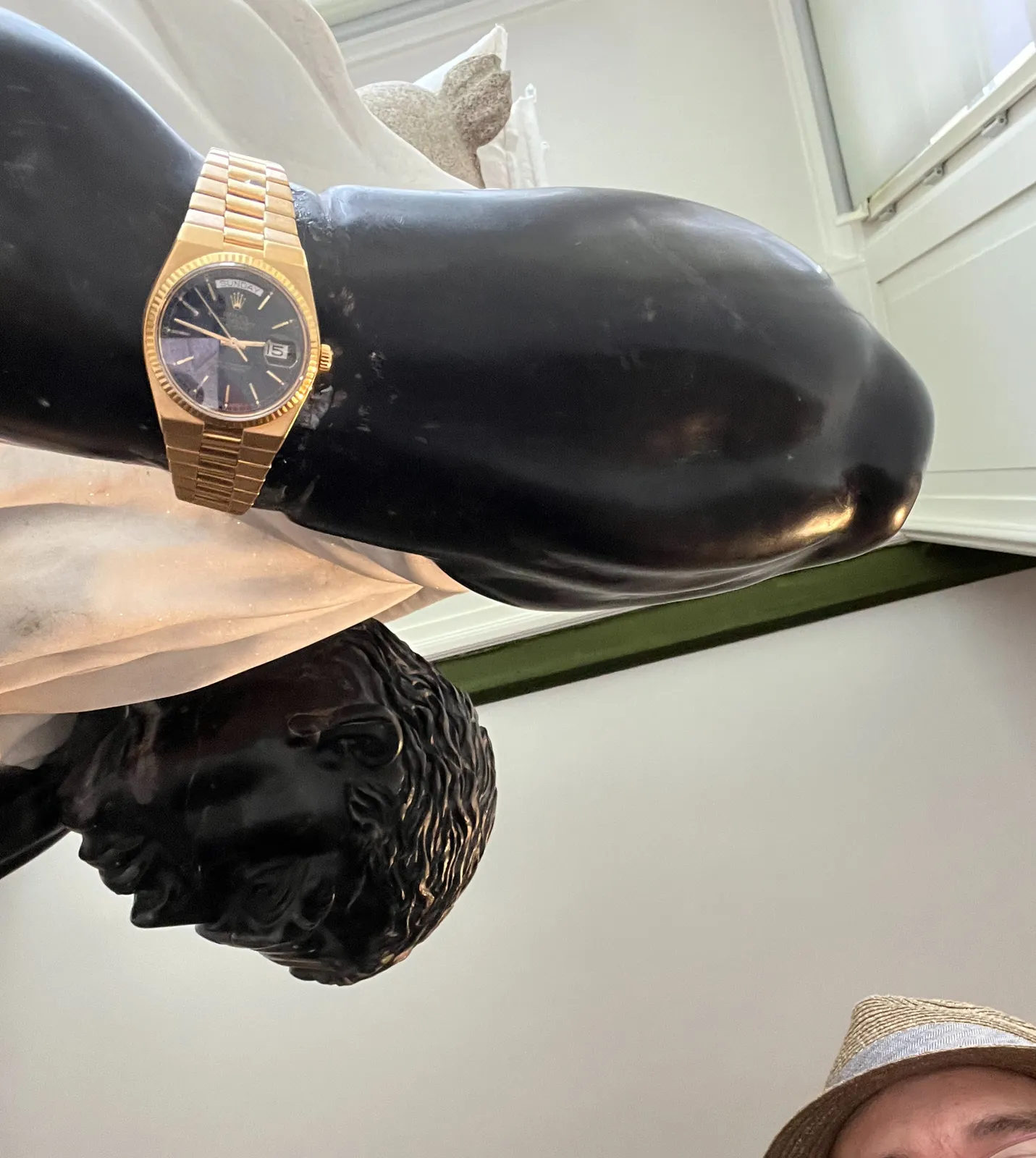 Looking up at a gold wrist watch that’s on the wrist of a black marble statue. The hat and the eye brow of the photographer can be seen. The watch points at 13 minutes to 3.