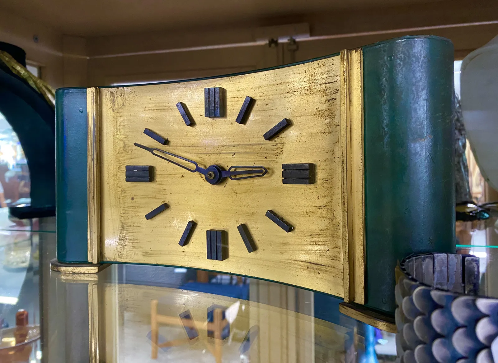 An old shelf clock. It’s curved inwards. The face is painted gold, and its sides are painted in a dark blueish green. It points at 12 minutes to 3.