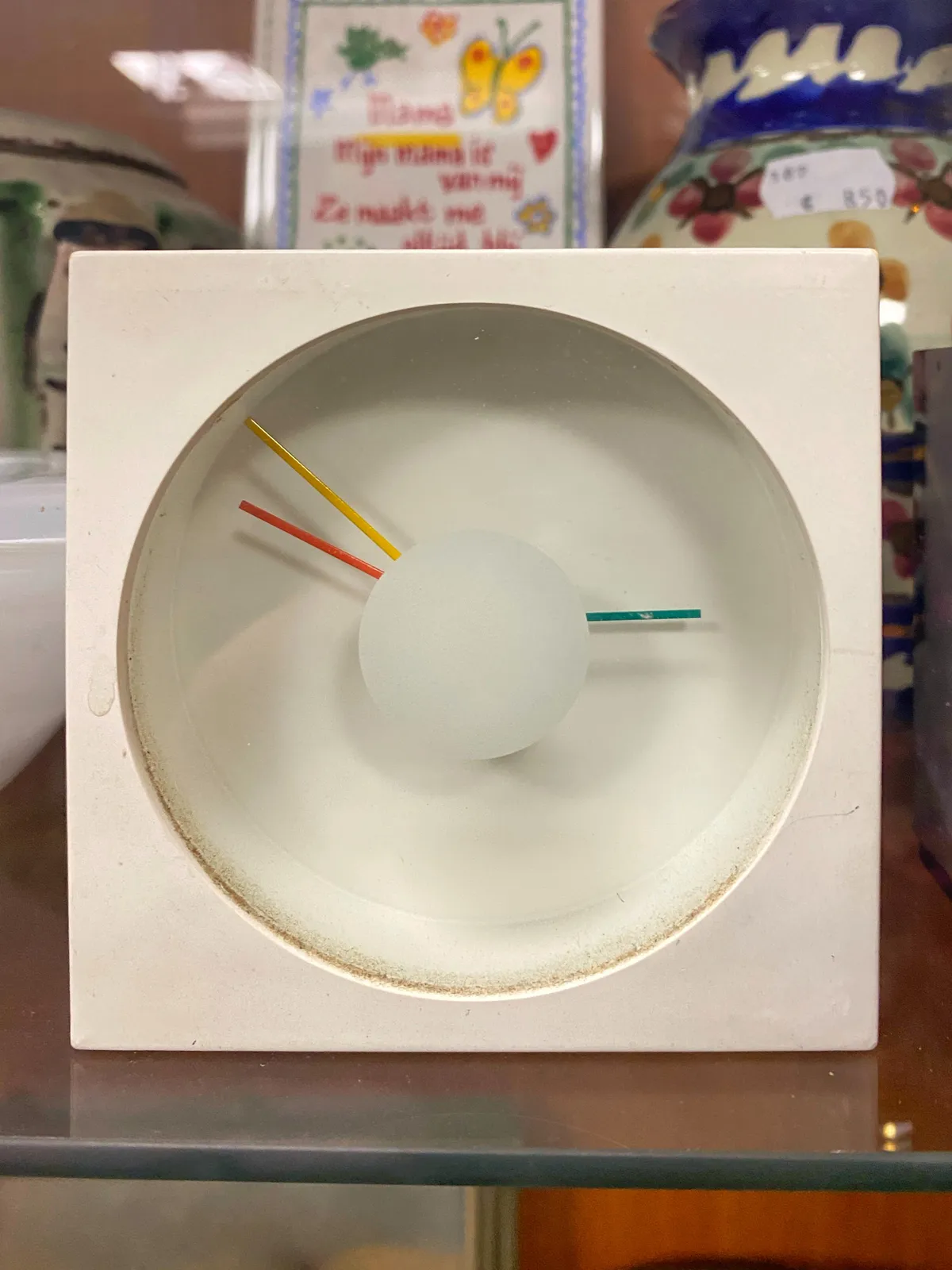 A rather minimalist clock. It’s all white, with no hour or minute indicators. It has a green hour hand, and it has two more hands. It’s unclear which one is the minute hand. Either the yellow one or the red one. So it’s either 12 minutes to 3, or 10 minutes to 3.
