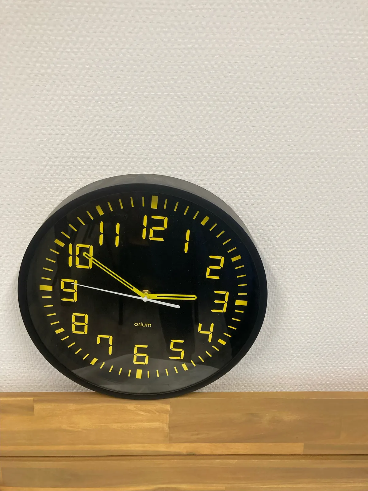 A black, modern wall clock, standing on the floor, a but skewed. The hour indicators are printed with so called, seven segment digits, which is very common for digital clocks, but rather uncommon for a non-digital clock like this one. The hands point at ten minutes to three.