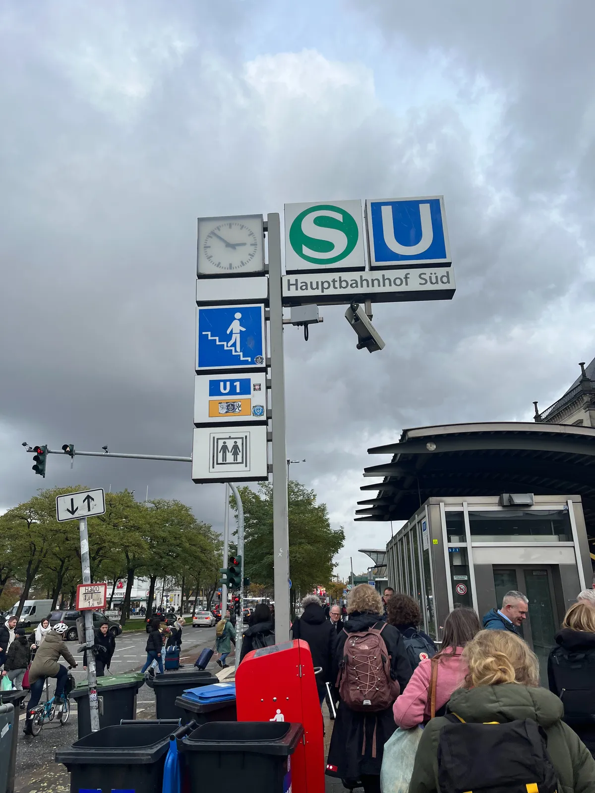 Many people walking towards metro station Hauptbahnhof Süd. On the pole which indicates the type of transport that’s leaving from this station there’s also a clock. It shows the time as 8 minutes to 3.