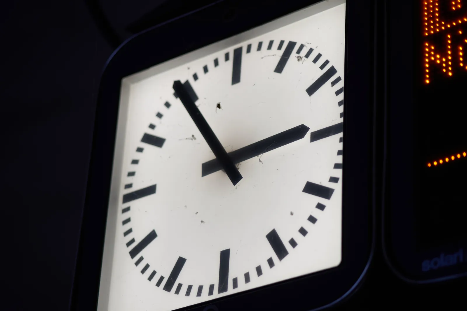 A dark image with a lit modernist clock, probably on a train station. The clock points at 6 minutes to 3.