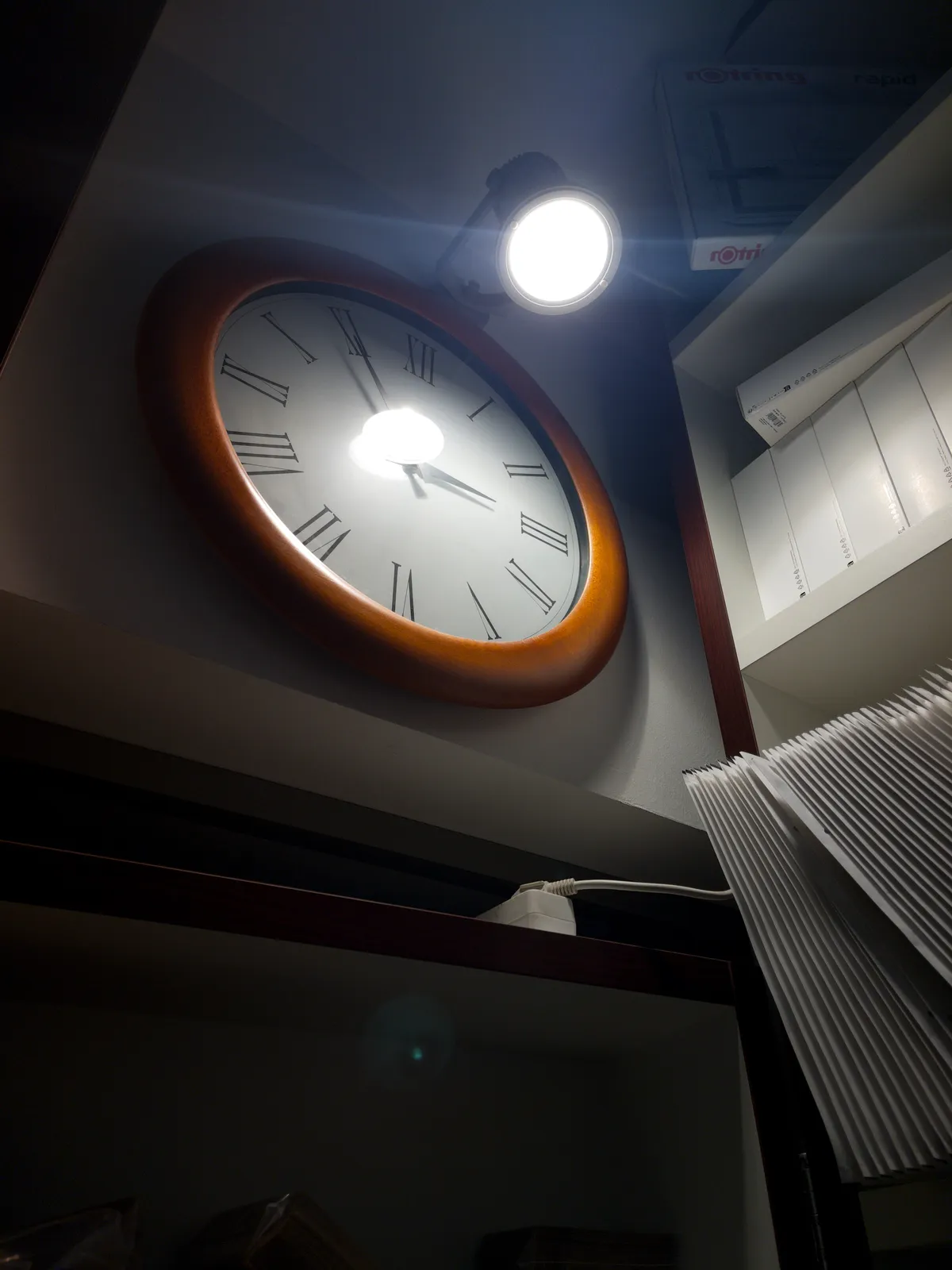 A clock up on a wall, next to a shelf with envelopes and boxes. There’s a lamp in the ceiling,  shining into our face. The clock points at 5 to 3.