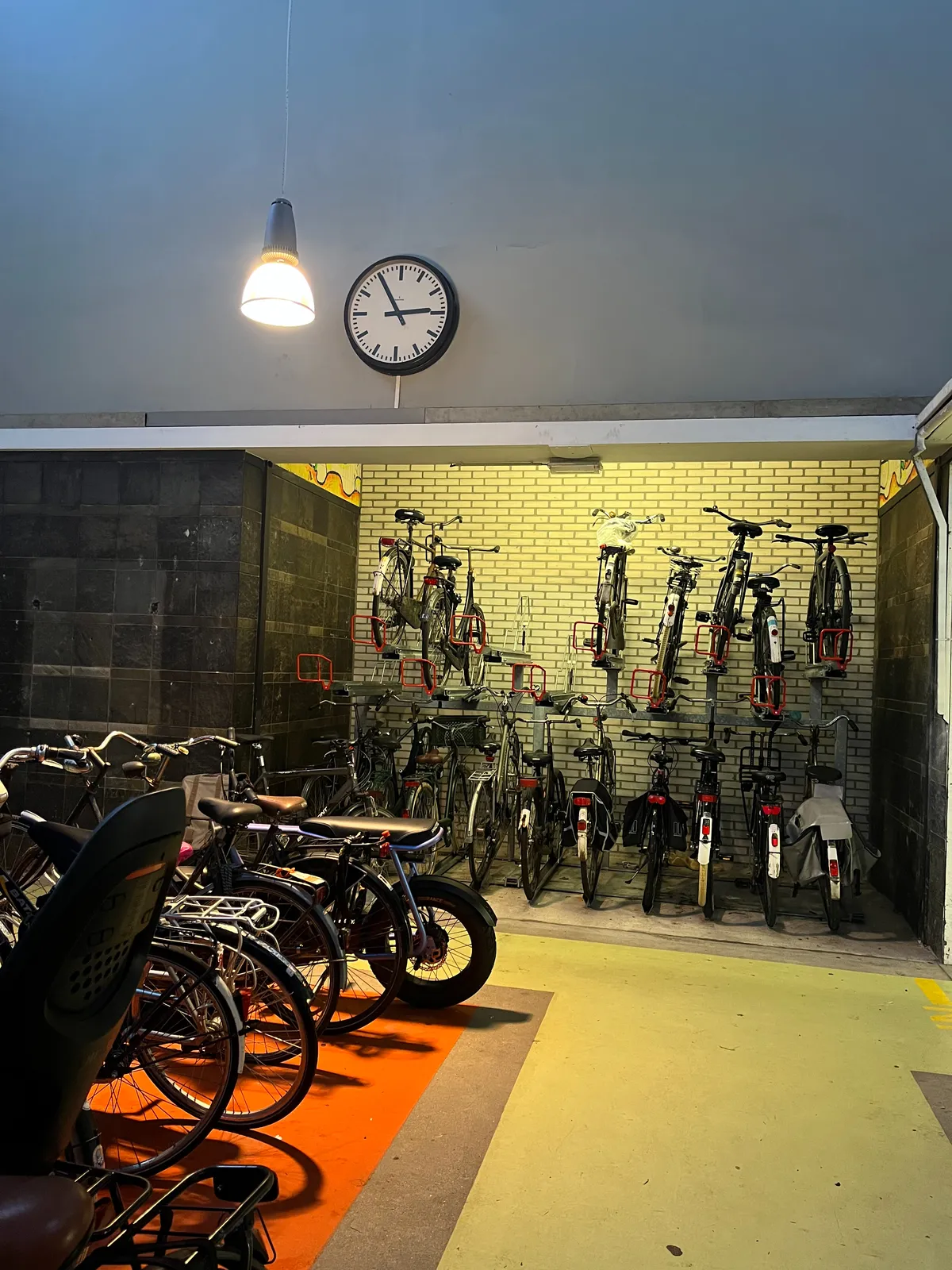 A clock attached to a wall in a pretty full bike parking. The clock points at 5 minutes to three.
