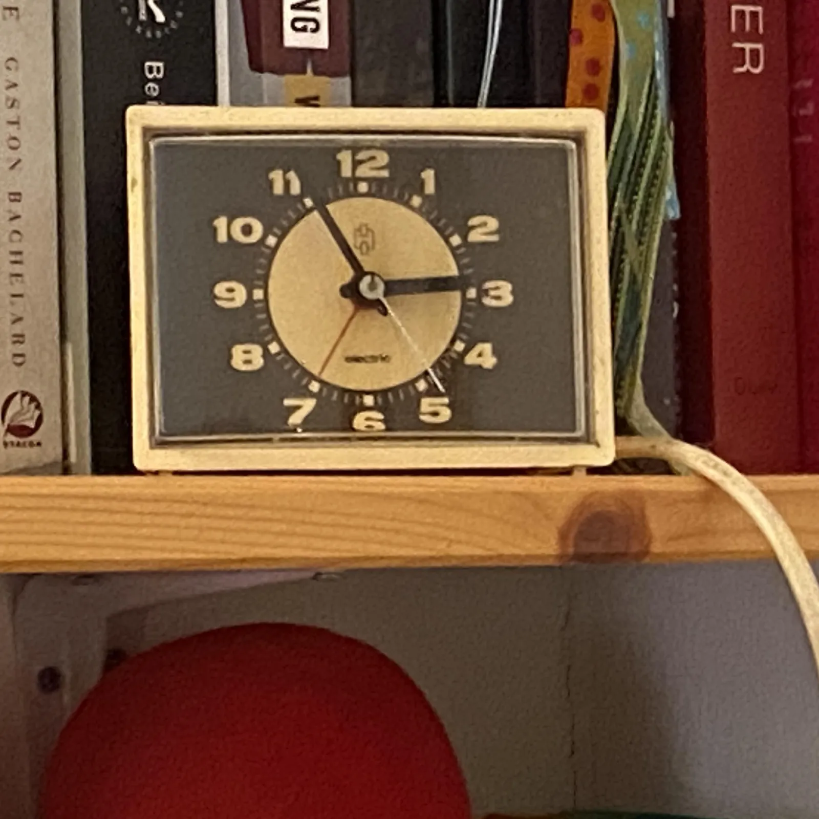 An alarm clock on a book shelf. It points at 4 minutes to 3.