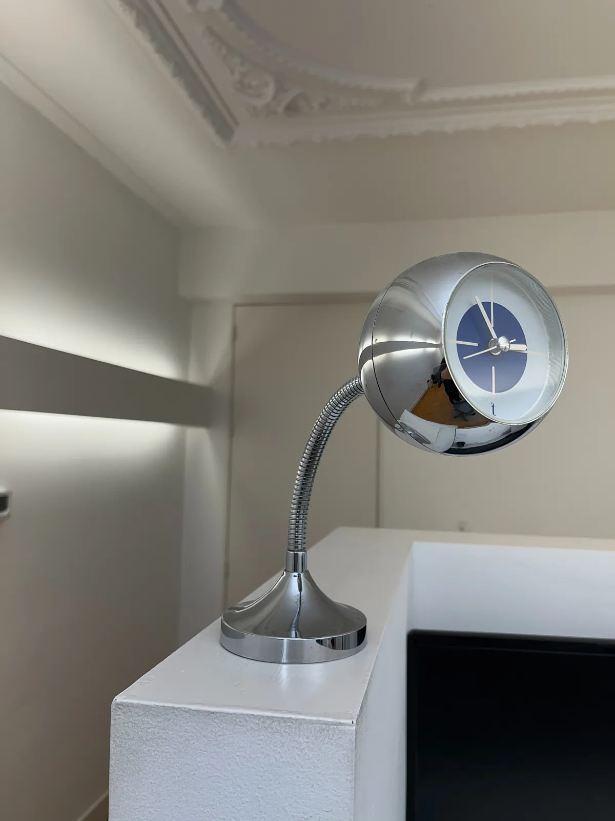 A design clock in a classical yet modern room. The clock is made from a shiny metal sphere on a bent tube. It points at 4 minutes to 3. 