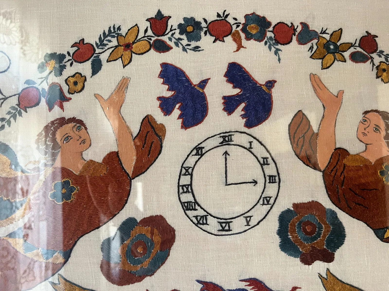 A detail of a piece of embroidery. There are two angels, two birds, and many flowers on it, surrounding a clock that points at three o’clock