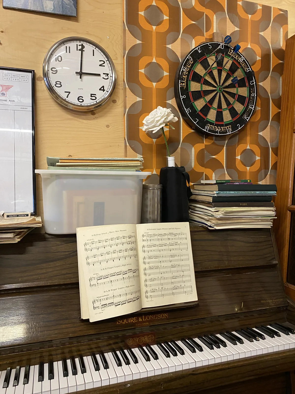 A piano, a darts board, some other stuff and a clock. On the clock it is three o’clock.