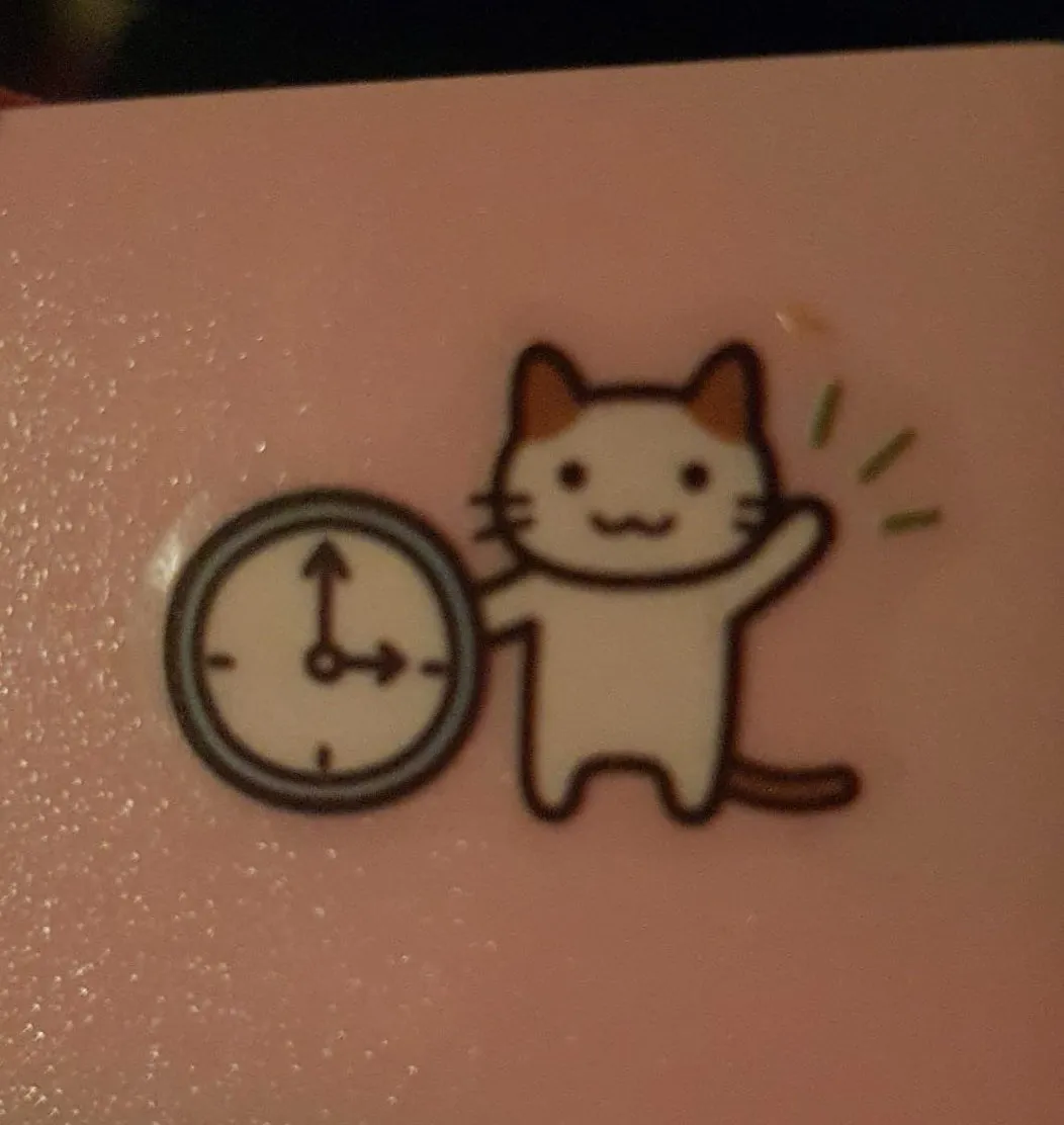 A blurry sticker with a cartoon drawing of a happy, standing cat, who’s waving at us. The cat leans against a clock that points at exactly three o’clock.