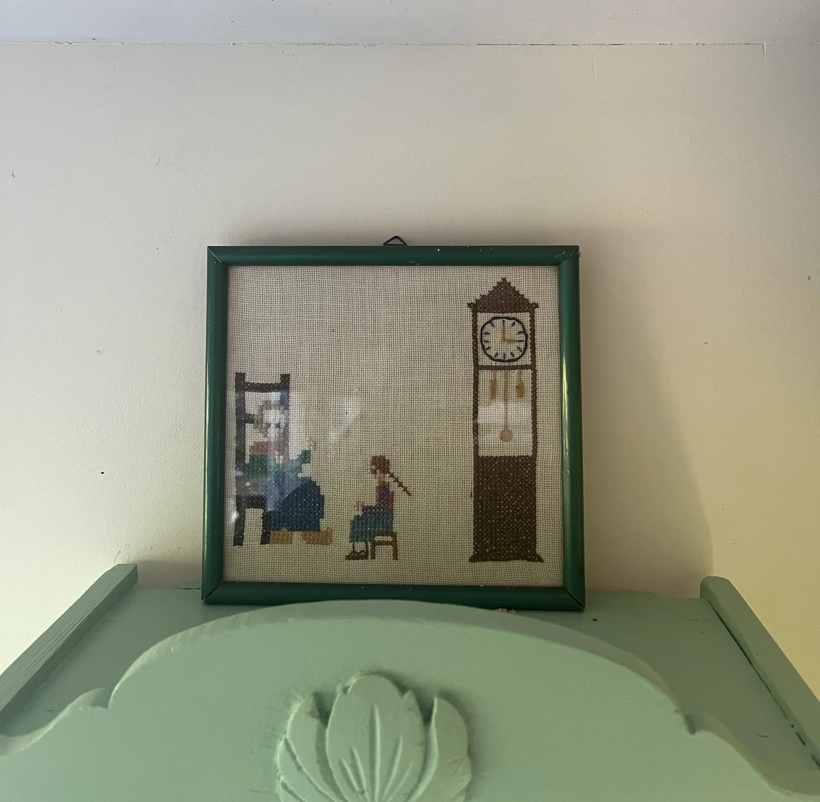 A framed piece of embroidery, with a scene of a child sitting next to a grandmother, and next to a standing clock. This clock points at three o’clock.