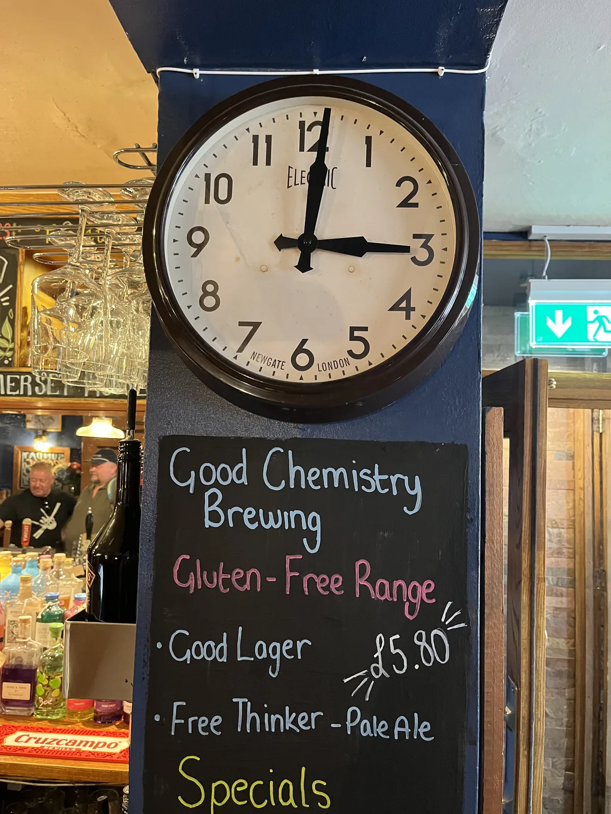 There’s a chalkboard which promotes gluten - free range beers from Good Chemistry Brewing, with names like Good Lager and Free Thinker Pale Ale, both for sale for five pound eighty. Above this board there’s a wall clock which points at one minute past three.
