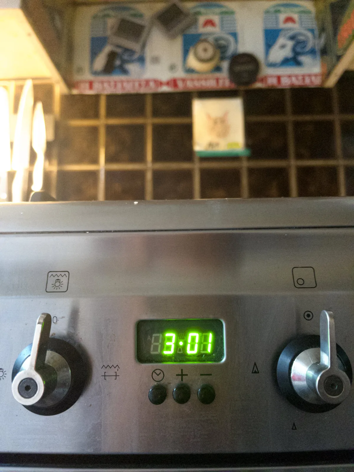 In the background, blurry, are some kitchen knives and some kitchen timers attached to a colourful wall. In the foreground are two knobs of a gas stove. Between these knobs there’s a digital clock that says that it is one minute past three.