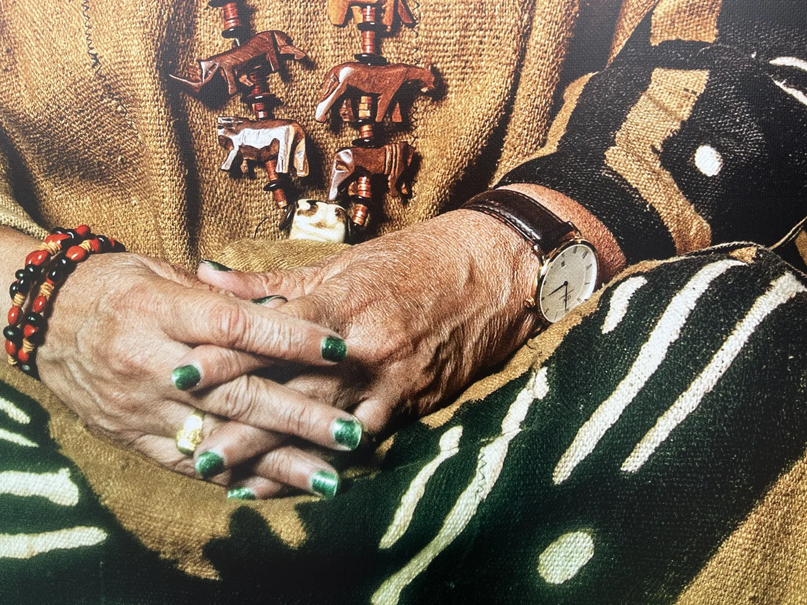 A detail from a larger picture. On this picture are the hands of an older person, with nice green nail polish. This person wears a brown, black white jersey, and a necklace made from wooden rhinos, tigers, and other animals. They also wear a wristwatch, which points at one minute past three.  