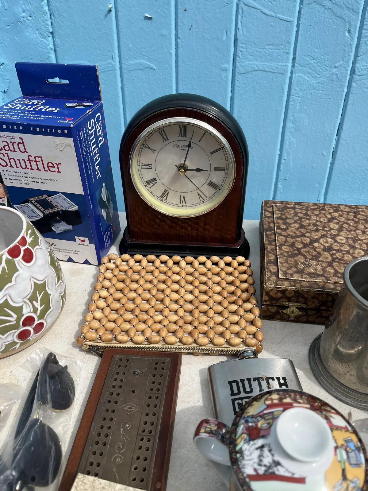 All kinds of stuff for sale on a table in a thrift store. A card shuffler, some sun glasses, and a box, among others. There’s also a mantel clock, which points at two minutes past three.