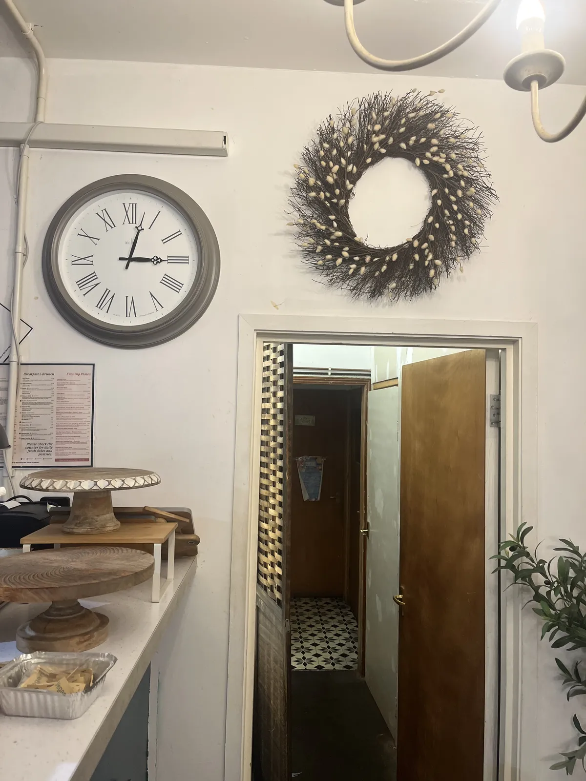 There are round plates, usually used for displaying cakes and such, but now they are empty. There’s a door which seems to lead to the toilets, there’s a round decoration made from twigs on the wall, and there’s a wall clock which points at 2 minutes past three. 