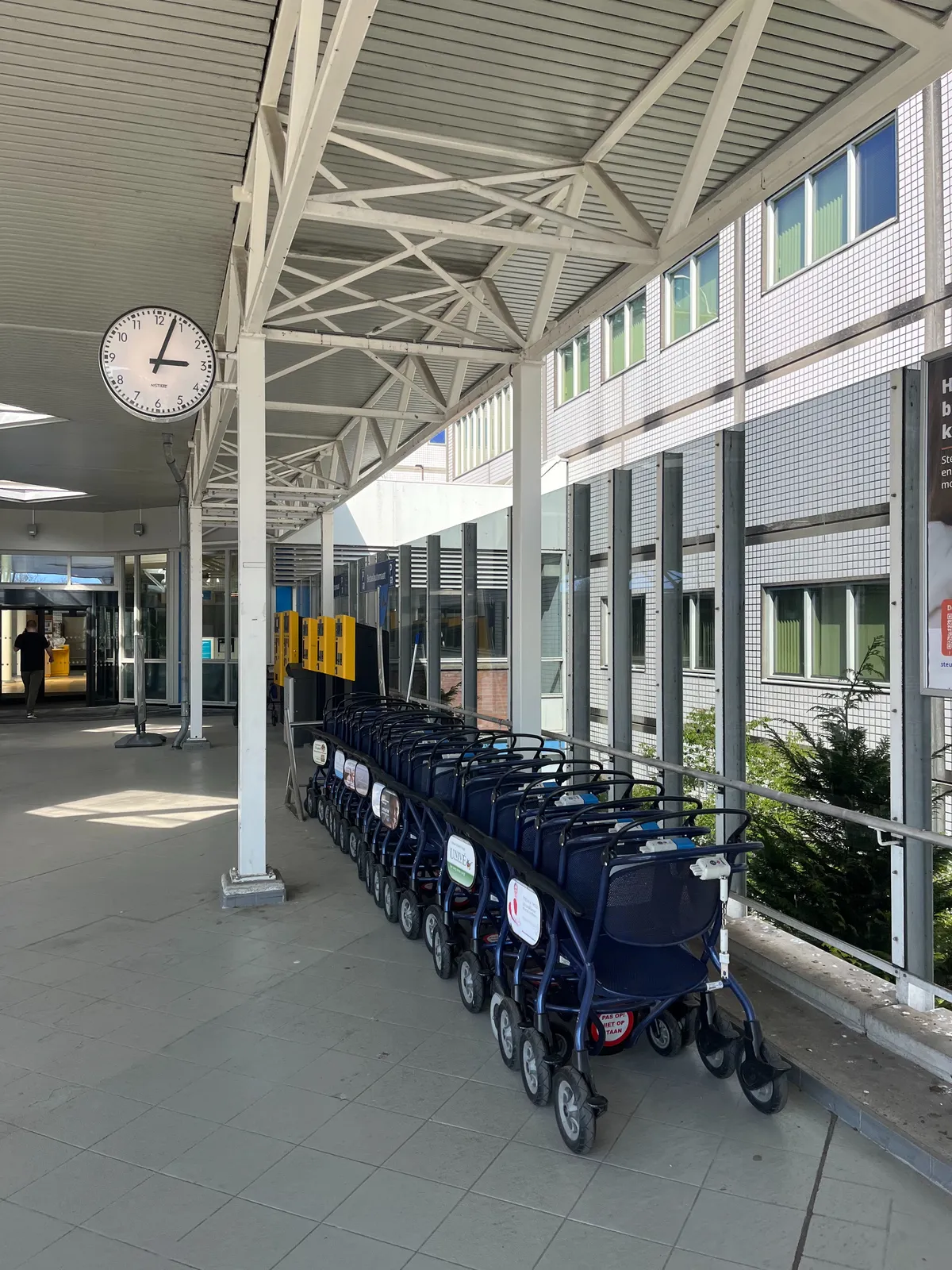 The entrance to a white building. Below a construction there is a row of parked, blue wheelchairs. Attached to the construction there’s a clock that points at 3 minutes past 3.