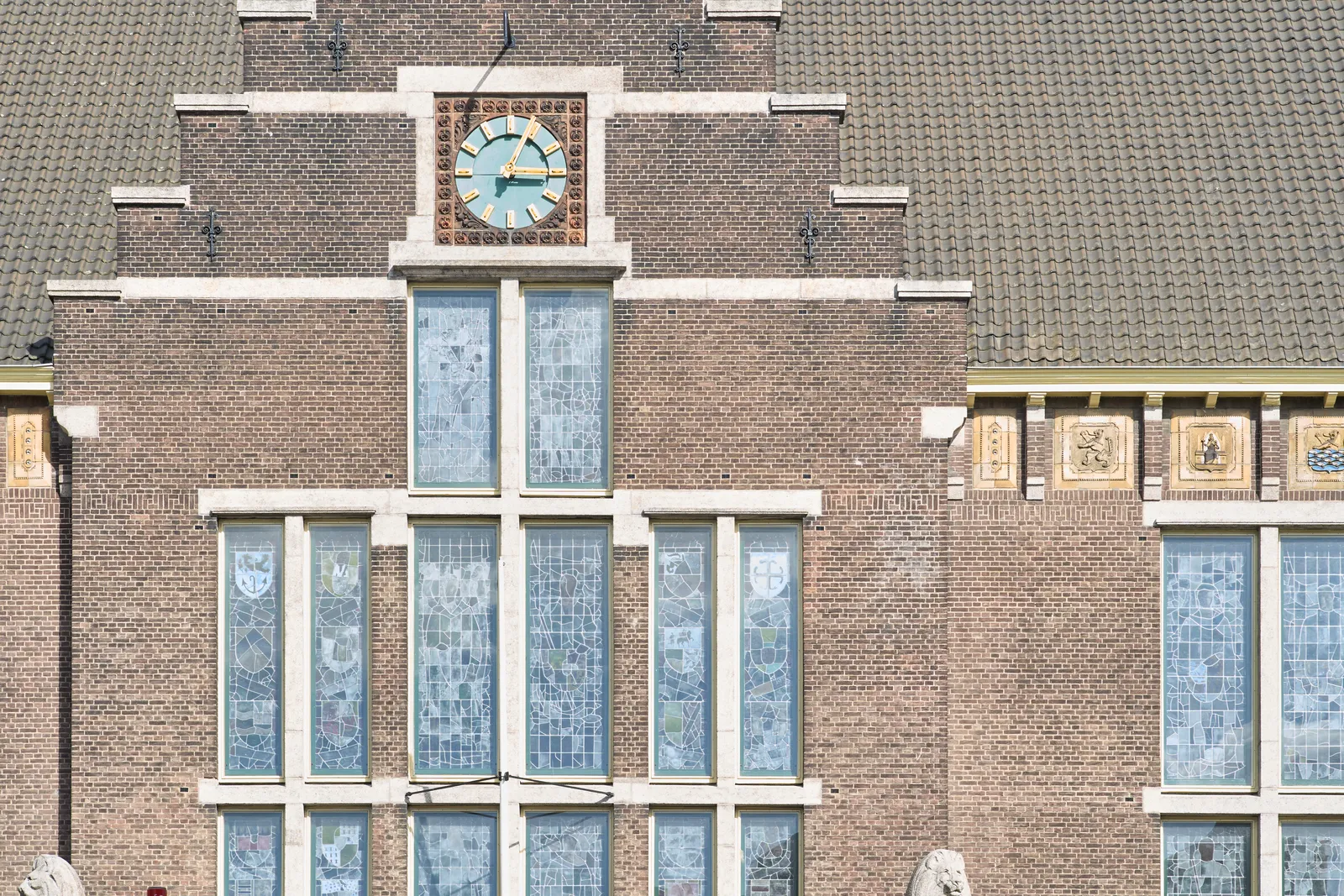 A clock built into a stepped facade of a medieval looking building, with ornamented glass-in-lead windows. The clock points at 4 minutes past three.